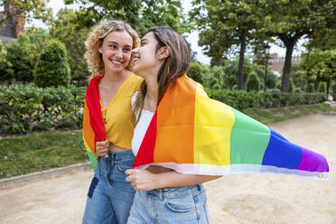 Lächelndes lesbisches junges Paar mit Regenbogenfahne, das im Park spazieren geht - WPEF06907