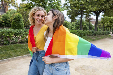 Smiling lesbian young couple covered with rainbow flag walking in park - WPEF06907