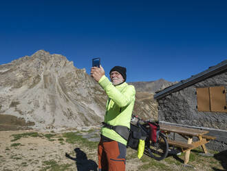 Ein älterer Mann macht ein Selfie mit seinem Mobiltelefon vor einem Berg unter blauem Himmel im Vanoise-Nationalpark, Frankreich - LAF02806