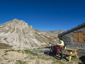 Älterer Mann mit Flasche auf einer Bank vor einem Mountainbike sitzend, Vanoise-Nationalpark, Frankreich - LAF02807