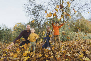 Glückliche Eltern mit verspielten Kindern, die Herbstblätter im Wald werfen - AANF00427