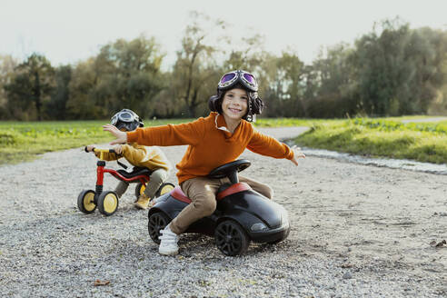 Lächelnder Junge sitzt mit ausgestreckten Armen auf einem Bobbycar mit seinem Bruder im Hintergrund - AANF00445