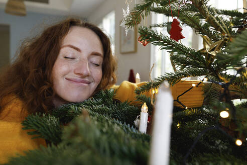 Lächelnde Frau mit geschlossenen Augen, die den Weihnachtsbaum zu Hause umarmt - TYF00640
