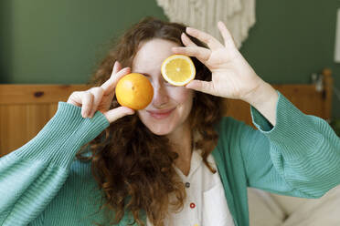 Smiling woman covering eyes with orange at home - TYF00650