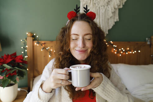 Smiling woman with eyes closed holding cup of hot chocolate on bed at home - TYF00656