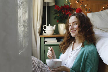 Smiling woman sitting with bowl by bed at home - TYF00665