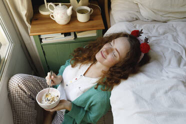 Smiling woman with eyes closed holding cereal bowl by bed at home - TYF00666