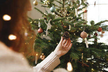 Hand einer Frau, die einen Tannenzapfen berührt, der zu Hause am Weihnachtsbaum hängt - TYF00680
