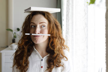 Woman balancing pen between nose and lips with book on head at home - TYF00685