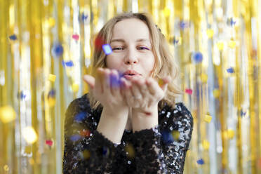 Woman wearing black dress blowing confetti in front of golden tinsel - ONAF00362
