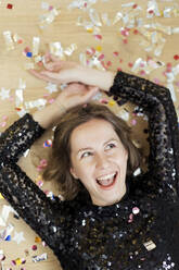 Happy woman wearing black dress lying on floor by confetti new year's party - ONAF00365