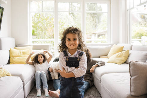 Glückliches Mädchen steht im Wohnzimmer mit Familie im Hintergrund zu Hause - MOEF04433