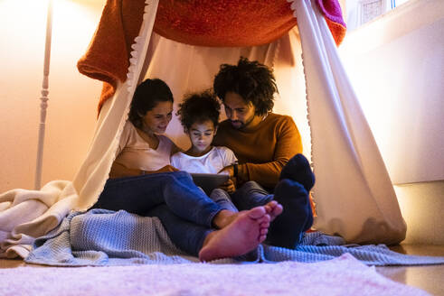 Mutter und Vater benutzen einen Tablet-PC, während ihre Tochter zu Hause im Zelt sitzt - MOEF04438