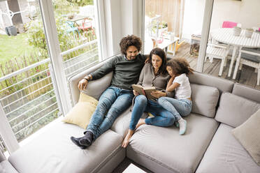 Father with mother reading book and daughter sitting on sofa at home - MOEF04440