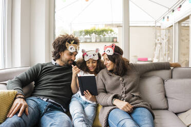 Smiling parents with daughter wearing eye masks and reading book on sofa at home - MOEF04442