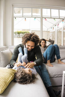 Glücklicher Vater, der seine Tochter auf dem Sofa kitzelt, während die Mutter im Hintergrund sitzt - MOEF04443