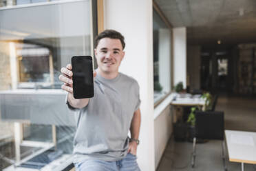 Lächelnder junger Geschäftsmann zeigt sein Smartphone, das auf der Fensterbank im Büro liegt - UUF28023