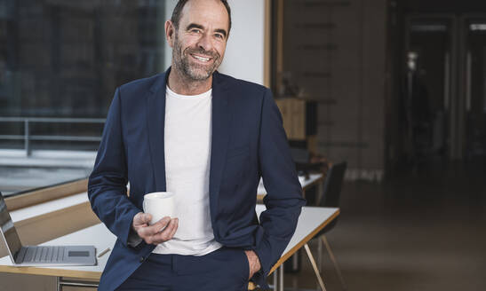 Glücklicher Geschäftsmann mit Kaffeetasse auf dem Schreibtisch im Büro - UUF28025