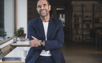 Happy businessman with arms crossed standing in office - UUF28032