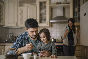 Vater und Sohn bereiten Tee zu, während die Mutter im Hintergrund ein Smartphone benutzt - ANAF00829