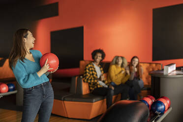 Glückliche junge Frau, die eine Kugel hält, mit Freunden im Hintergrund, die auf einer Bowlingbahn sitzen - JCCMF08761