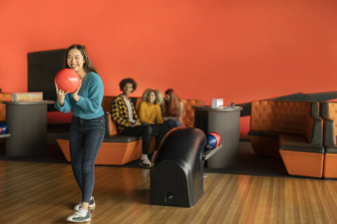 Lächelnde junge Frau beim Bowling mit Freunden im Hintergrund - JCCMF08762
