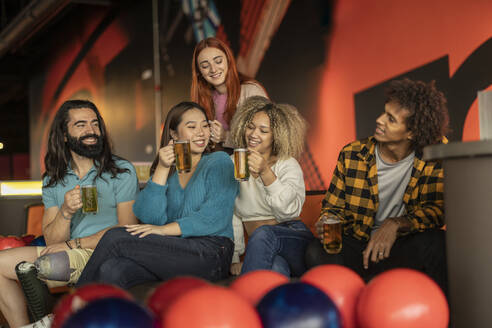Fröhliche, gemischtrassige Freunde sitzen mit einem Glas Bier auf einem Sofa in einer Bowlingbahn - JCCMF08795
