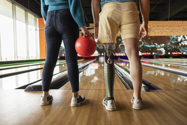 Frau, die eine Kugel hält, mit einem Freund, der eine Prothese trägt, auf dem Hartholzboden einer Bowlingbahn - JCCMF08806