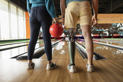 Frau, die eine Kugel hält, mit einem Freund, der eine Prothese trägt, auf dem Hartholzboden einer Bowlingbahn - JCCMF08806