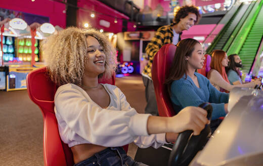 Lächelnde junge Frau mit Freunden, die einen Fahrsimulator in einer Spielhalle genießen - JCCMF08827