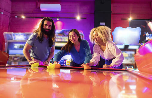 Multiracial friends having fun playing air hockey at amusement arcade - JCCMF08833