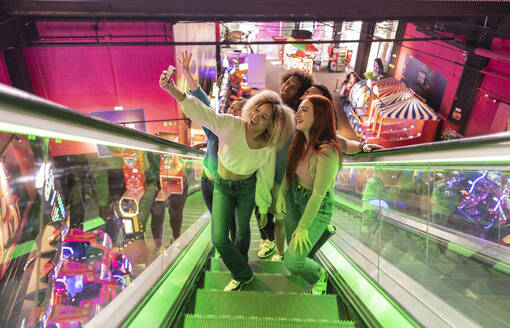 Multirassische Freunde, die von der Rolltreppe hochfahren und ein Selfie mit ihrem Smartphone in einer Spielhalle machen - JCCMF08841