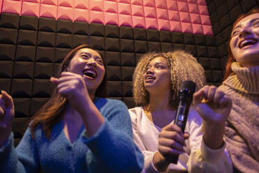 Happy friends enjoying holding microphone at amusement arcade - JCCMF08848