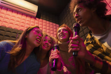 Multiracial friends singing karaoke through microphones at amusement arcade - JCCMF08850