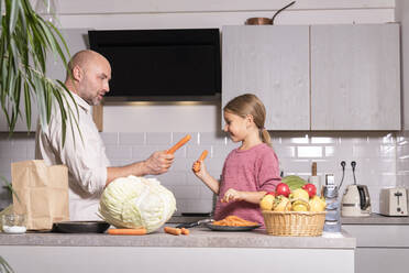 Father and daughter having fun with carrots in kitchen at home - KMKF01912