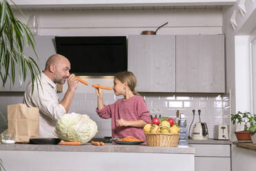 Father and daughter having fun with carrots in kitchen at home - KMKF01913