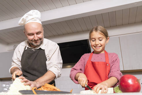 Vater und Tochter mit Schürzen bei der Zubereitung einer gesunden Mahlzeit in der Küche zu Hause - KMKF01923