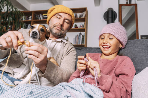 Vater und glückliche Tochter mit Hund stricken auf der Couch zu Hause zusammen - KMKF01930