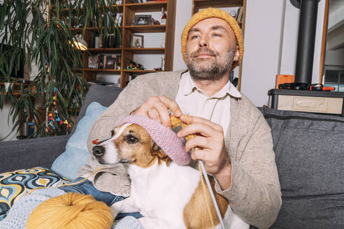 Mann mit Hund strickend auf der Couch zu Hause - KMKF01931