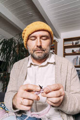 Man in warm clothing knitting on couch at home - KMKF01936