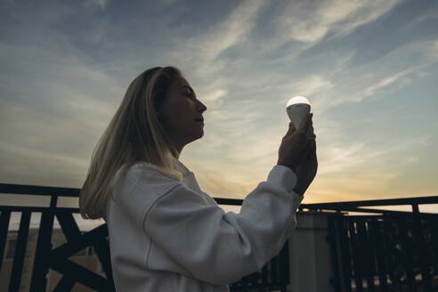 Frau hält LED-Glühbirne stehend unter Himmel bei Sonnenuntergang - SIF00651