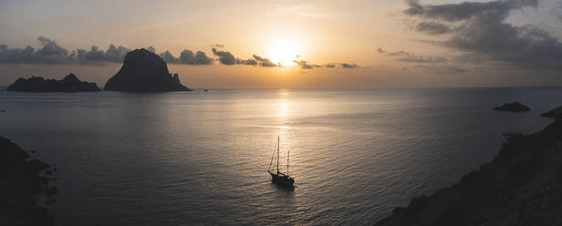 Spanien, Balearen, Segelboot schwimmt in der Nähe des Ufers bei Sonnenuntergang mit der Insel Es Vedra im Hintergrund - JAQF01162