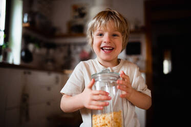 Porträt eines niedlichen kleinen Jungen, der einen Behälter mit Cornflakes zu Hause hält und in die Kamera schaut. - HPIF05596