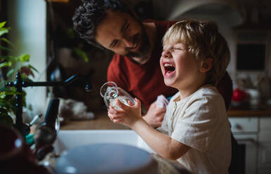 A small laughing boy with father having fun when washing dishes indoors at home, daily chores concept. - HPIF05607