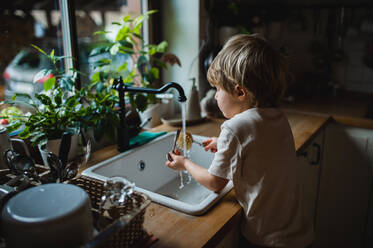 Ein kleiner Junge, der eine umweltfreundliche Bürste benutzt, um das Geschirr zu Hause zu waschen, ein Konzept für die tägliche Arbeit. - HPIF05610