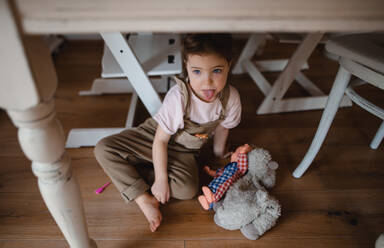 A portrait of cute small girl sitting on floor under table indoors at home, sticking tongue out. - HPIF05618