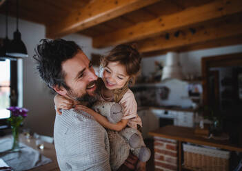 A mature father with small daughter standing indoors at home, holding and hugging. - HPIF05619
