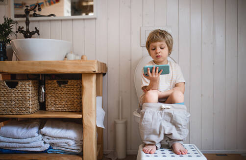 Ein Porträt eines süßen kleinen Jungen, der zu Hause auf der Toilette sitzt und sein Smartphone benutzt. - HPIF05662