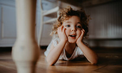 A portrait of cute small girl lying on floor under table indoors at home, looking away. - HPIF05674