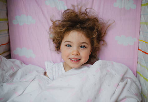 A top view of happy little girl lying in bed indoors at home - HPIF05676